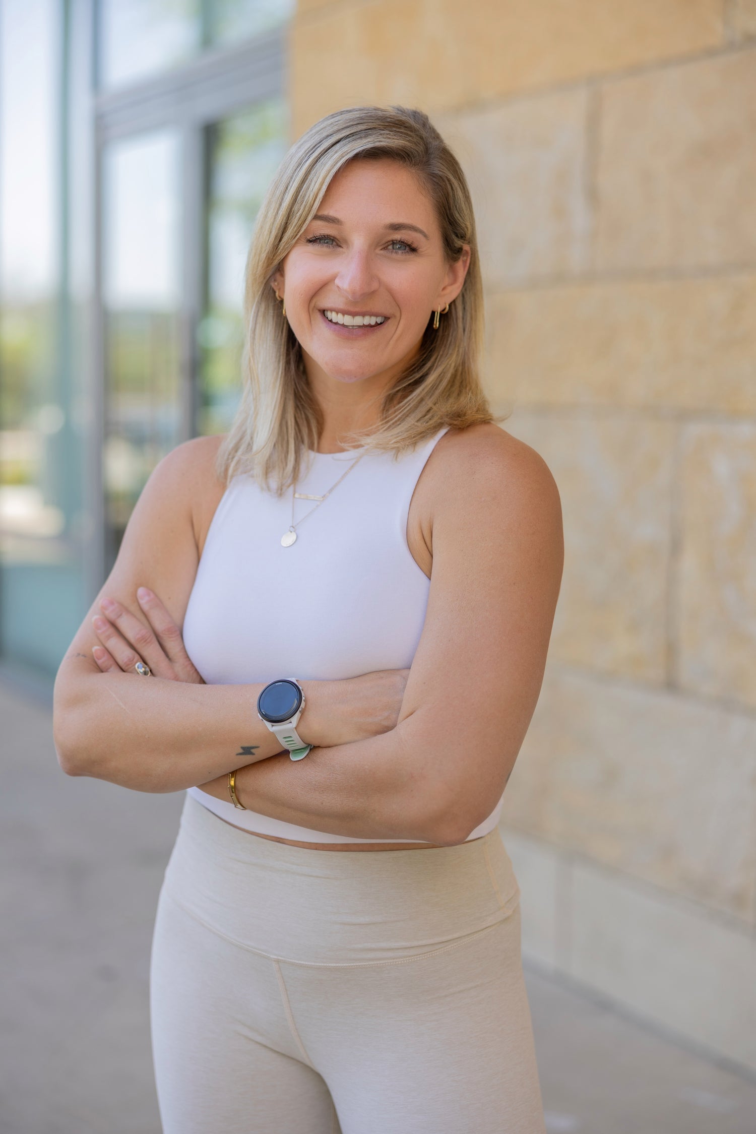Chiara Adin Moore. A smiling blonde woman stands outdoors with her arms crossed, wearing a white sleeveless top and light beige leggings. She is positioned in front of a beige stone wall and glass windows.