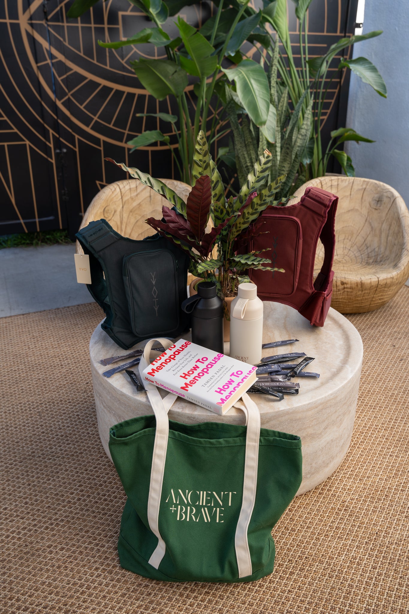 Green bag with 'Ancient Brave' branding on a round stone table with plants and bottles.